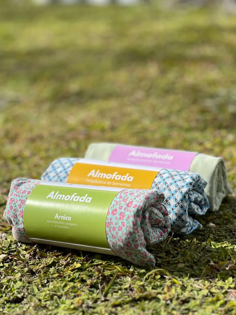 Três almofadas terapêuticas de sementes enroladas em tecido estampado, sobre a grama