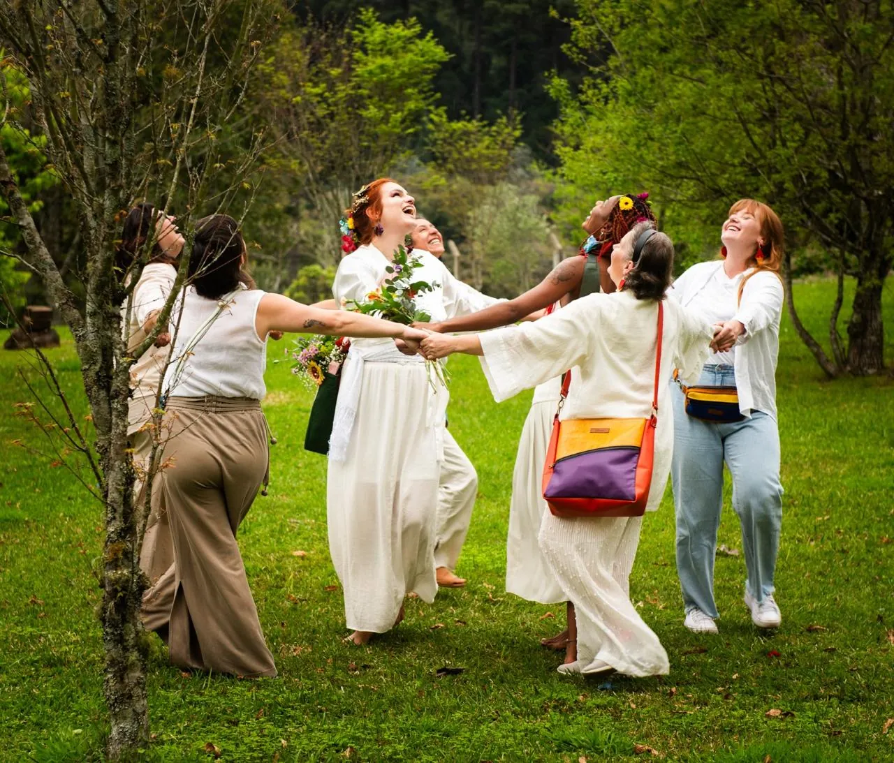 Mulheres vestidas de branco dançando em roda num gramado, com bolsas coloridas